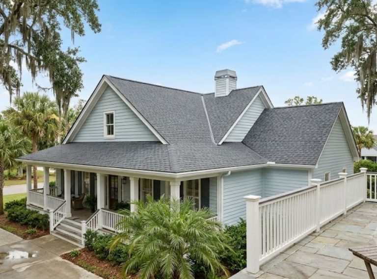 Southern-style house with light blue siding, complex dark roof, and covered porch in a mature garden.