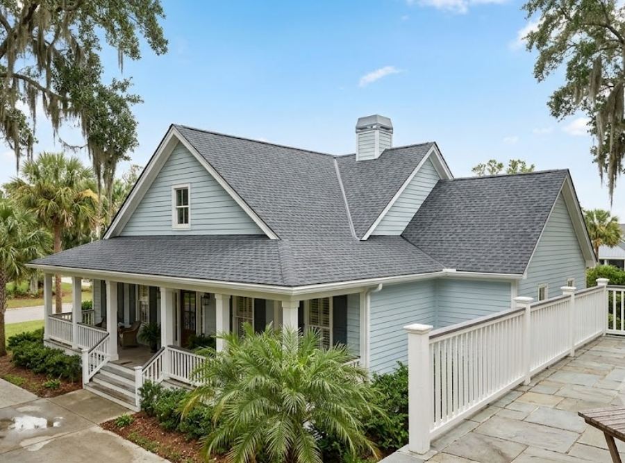 Southern-style house with light blue siding, complex dark roof, and covered porch in a mature garden.